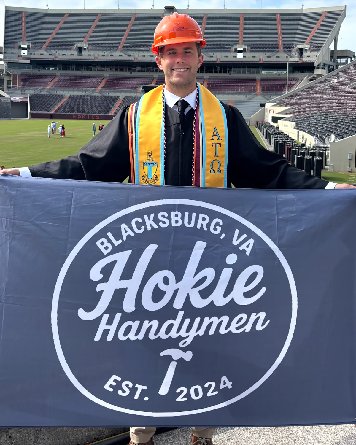 Robbie in graduation robes and an orange hard hat holding a Hokie Handymen banner at Virginia Tech